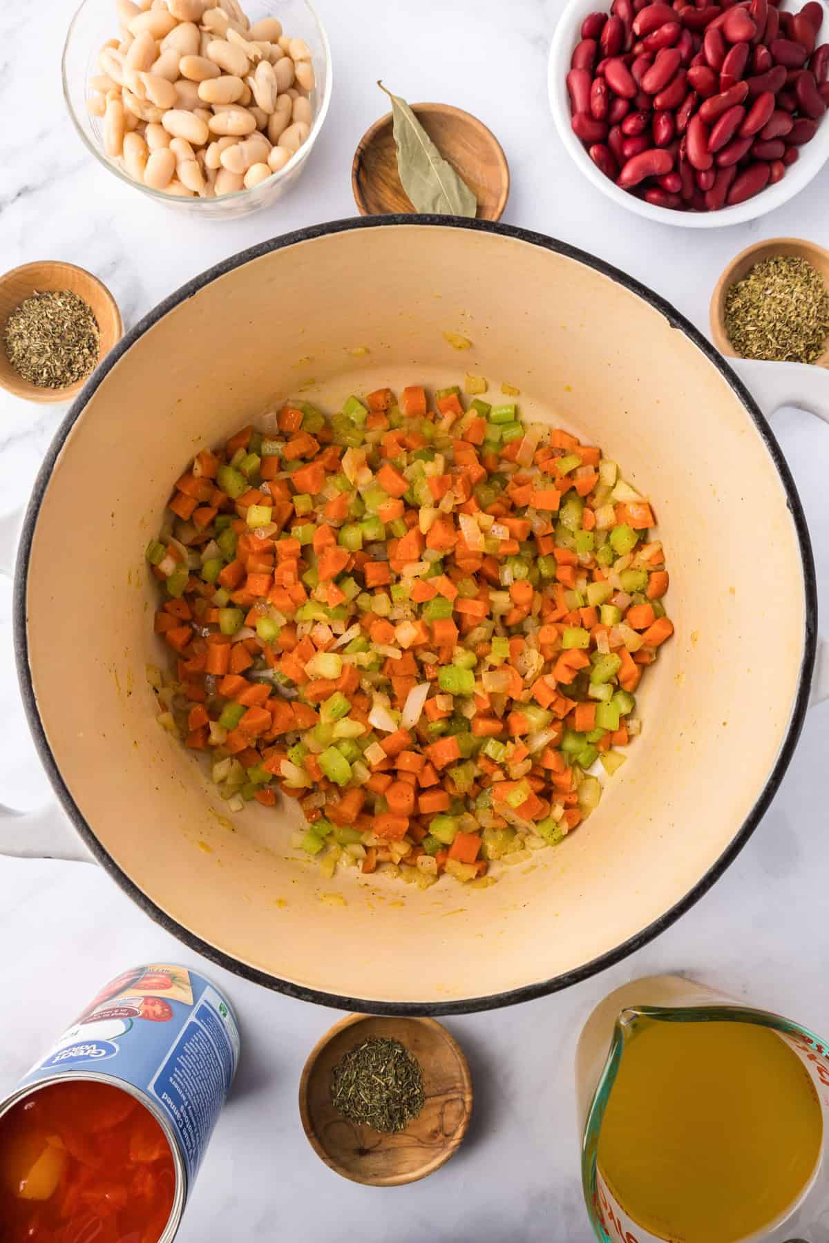 Chopped carrots, celery, and onion are sautéing in a large pot for a hearty minestrone soup, surrounded by bowls of beans, herbs, bay leaf, canned tomatoes, and a measuring cup of broth.