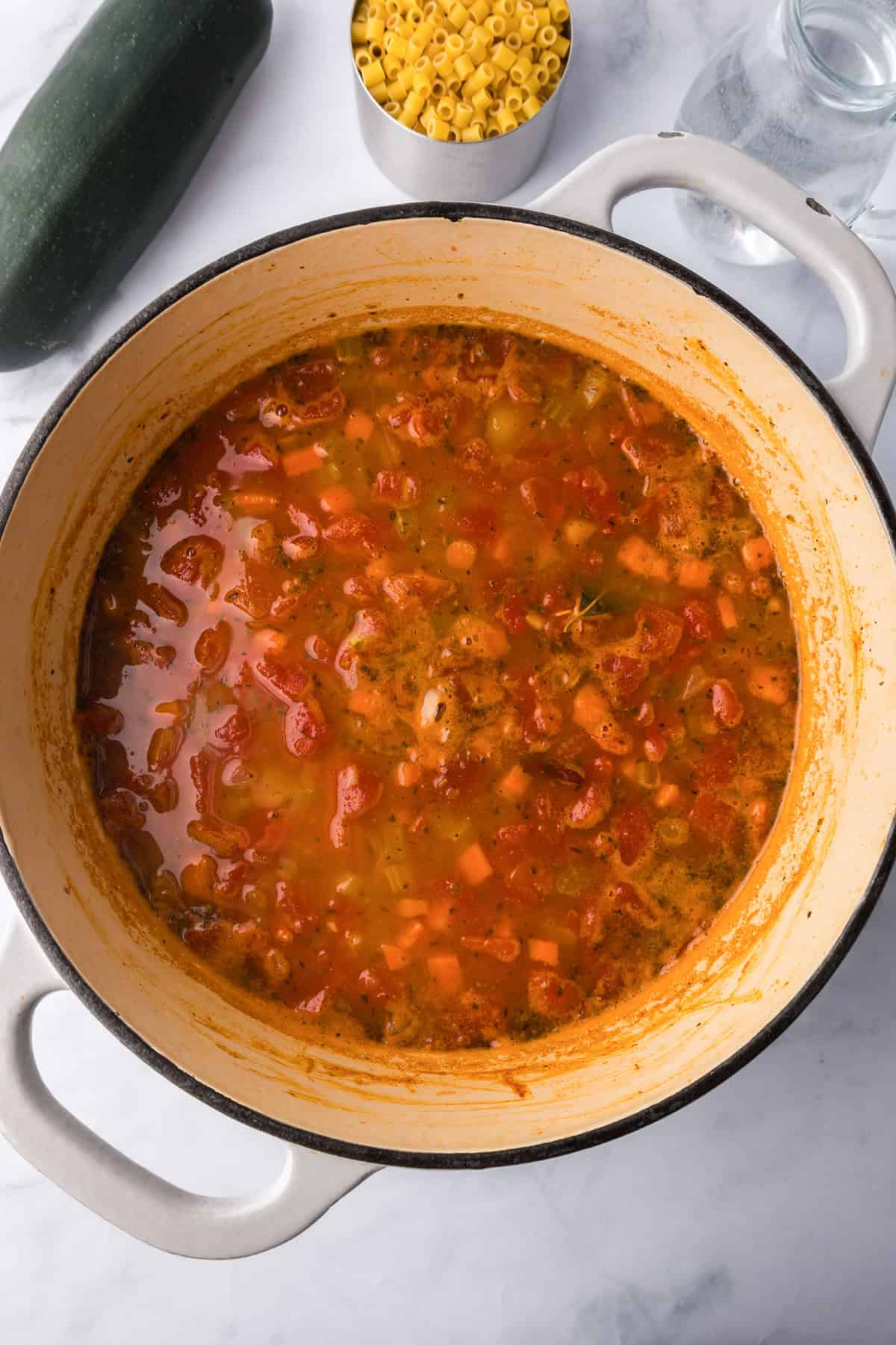 A large pot filled with hearty minestrone soup in a tomato-based broth, with diced vegetables visible. Nearby are a zucchini, a bowl of uncooked pasta, and a glass water bottle.