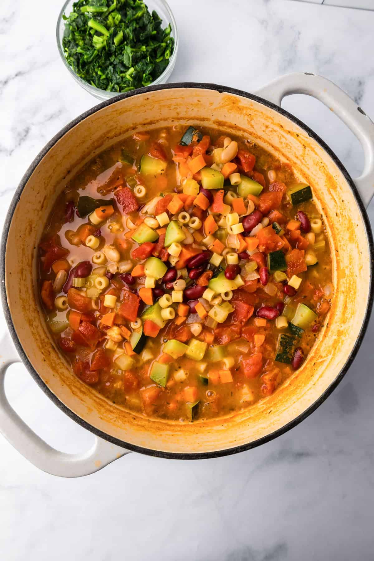 A pot of minestrone soup with ditalini pasta, beans, chopped zucchini, carrots, and tomatoes, served alongside a bowl of chopped greens.