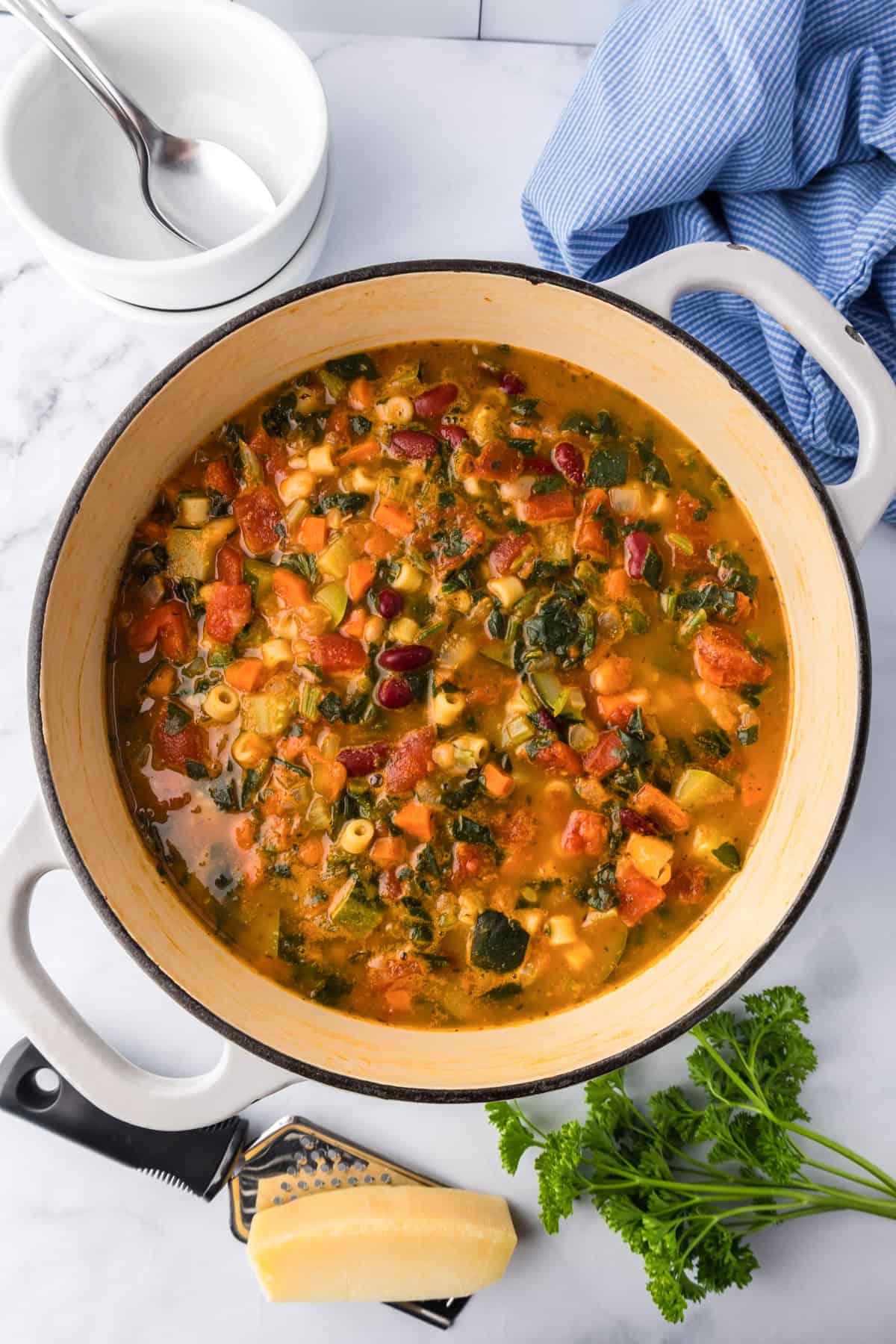 A pot of minestrone soup with pasta, beans, tomatoes, and greens sits on a white countertop beside a block of cheese, grater, parsley, stacked bowls, and a blue cloth.