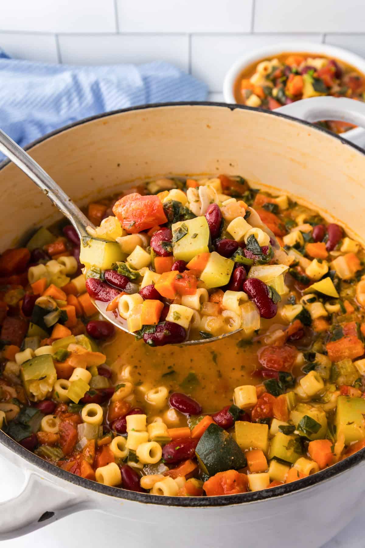 A ladle lifts a serving of hearty minestrone soup with ditalini pasta from a large pot. The soup contains carrots, zucchini, tomatoes, and kidney beans.