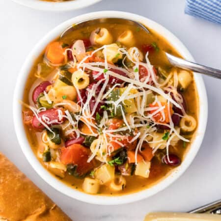 A bowl of minestrone soup with pasta, kidney beans, and diced vegetables, topped with grated cheese, sits on a white surface next to bread.