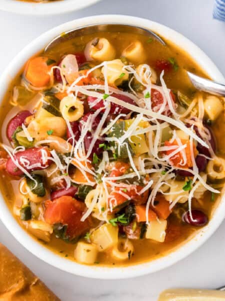 A bowl of minestrone soup with pasta, kidney beans, and diced vegetables, topped with grated cheese, sits on a white surface next to bread.