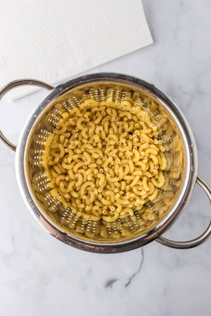 Cooked elbow macaroni noodles draining in a metal colander on a marble countertop, ready to be used in a creamy slow cooker mac and cheese recipe, with a paper towel nearby.