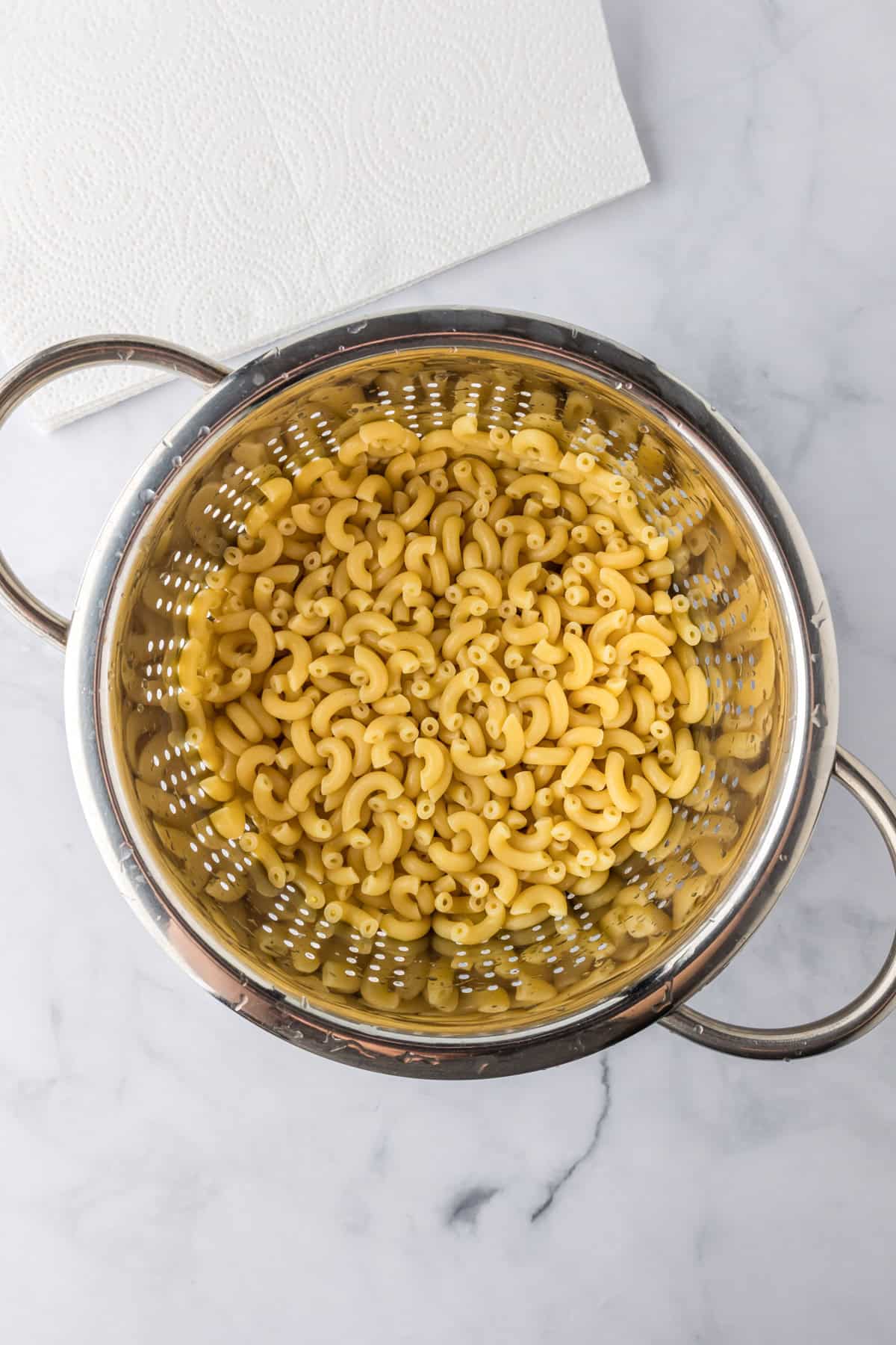 Cooked elbow macaroni noodles draining in a metal colander on a marble countertop, ready to be used in a creamy slow cooker mac and cheese recipe, with a paper towel nearby.
