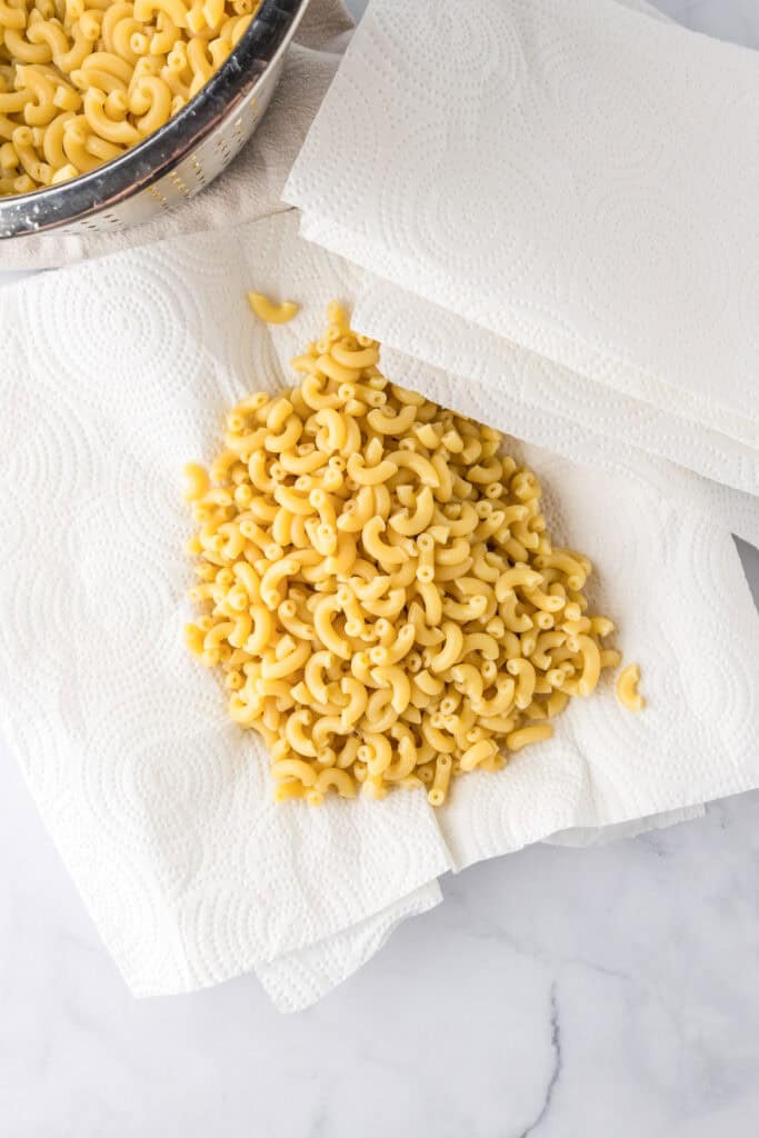 Cooked elbow macaroni, perfect for slow cooker mac and cheese, is spread out on paper towels to dry, with a colander and extra paper towels.