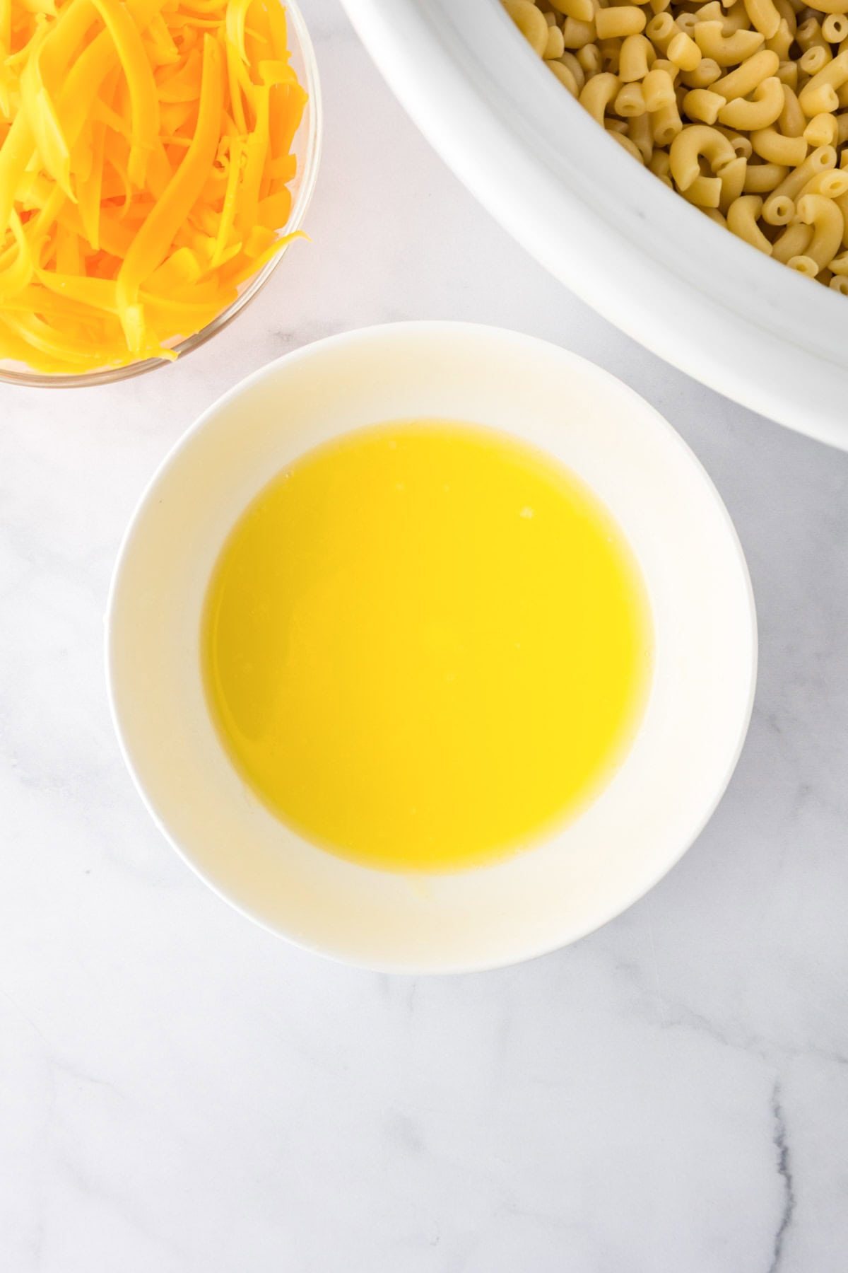 A bowl of melted butter sits on a marble surface next to shredded cheddar cheese and uncooked elbow macaroni—perfect ingredients for homemade slow cooker mac and cheese.