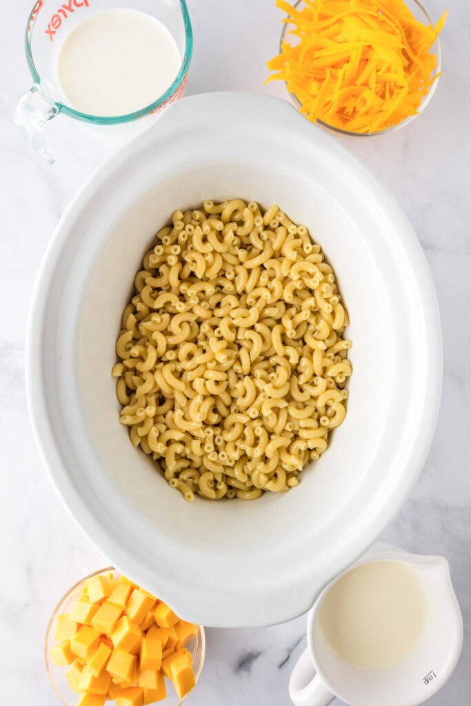 A slow cooker mac and cheese setup with dry elbow macaroni, shredded cheddar cheese, cheese cubes, a measuring cup of milk, and half-and-half arranged on a white surface.