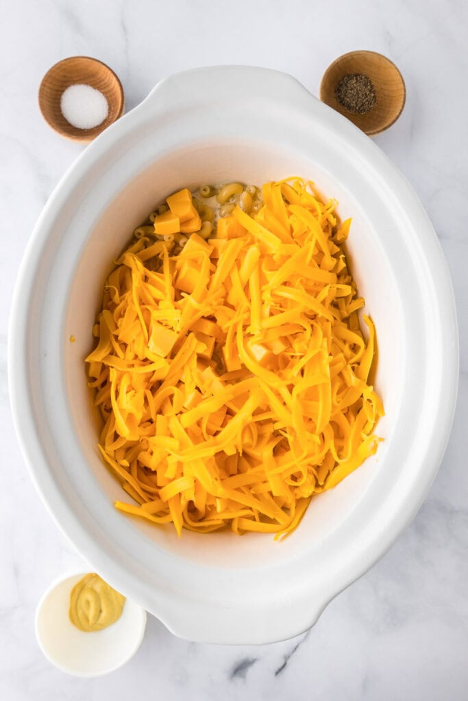 Shredded cheddar cheese, butter, and other ingredients for slow cooker mac and cheese are in a white slow cooker, with small bowls of salt, pepper, and mustard on a marble countertop.