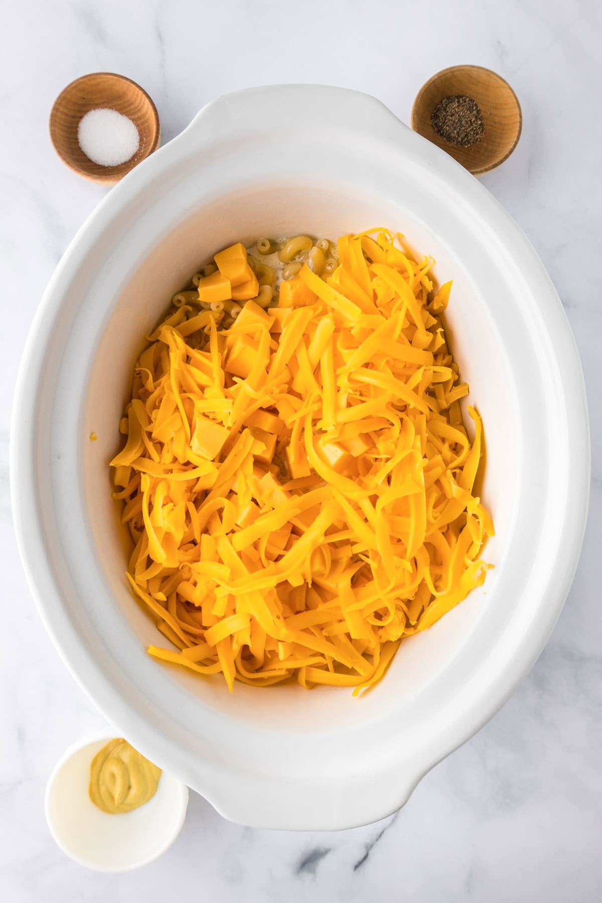 Shredded cheddar cheese, butter, and other ingredients for slow cooker mac and cheese are in a white slow cooker, with small bowls of salt, pepper, and mustard on a marble countertop.