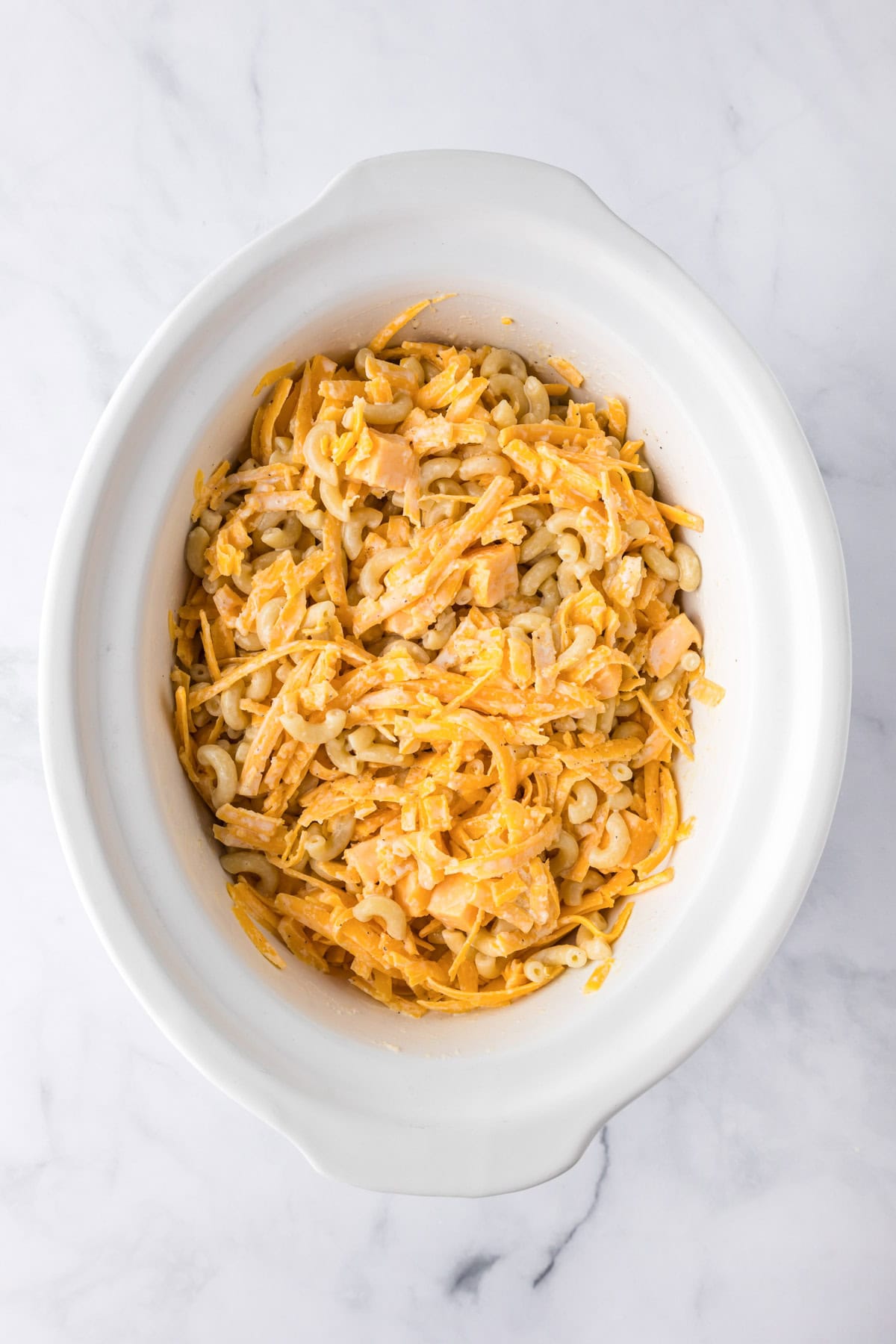 Oval white slow cooker base filled with uncooked macaroni, shredded cheddar cheese, and a creamy sauce on a marble surface, perfect for an easy slow cooker mac and cheese recipe.