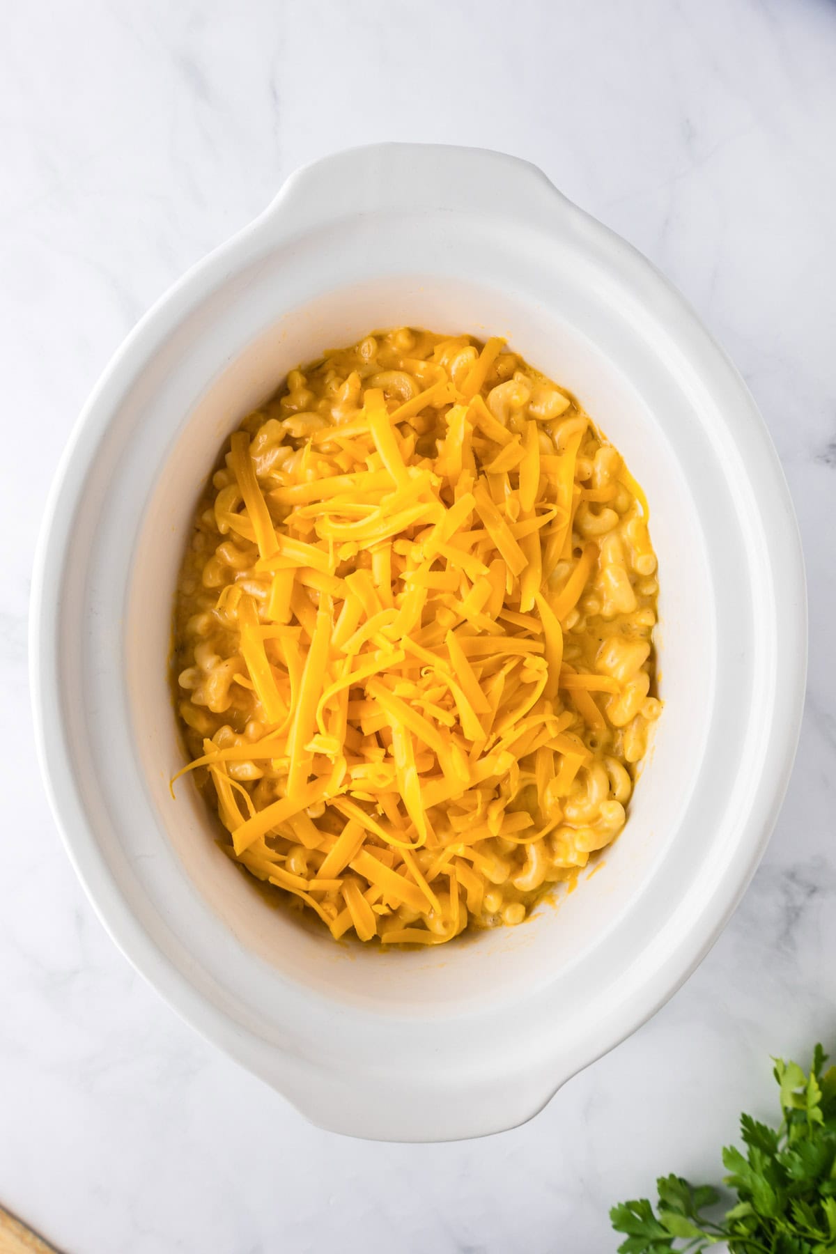 A white slow cooker filled with creamy slow cooker mac and cheese, topped with shredded cheddar cheese, sits on a marble surface.