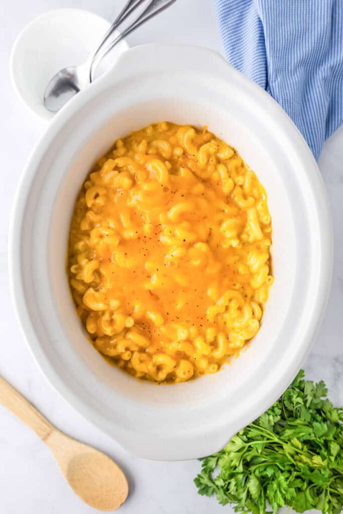 A slow cooker filled with creamy slow cooker mac and cheese sits on a white surface, surrounded by a wooden spoon, fresh parsley, and a blue striped cloth.