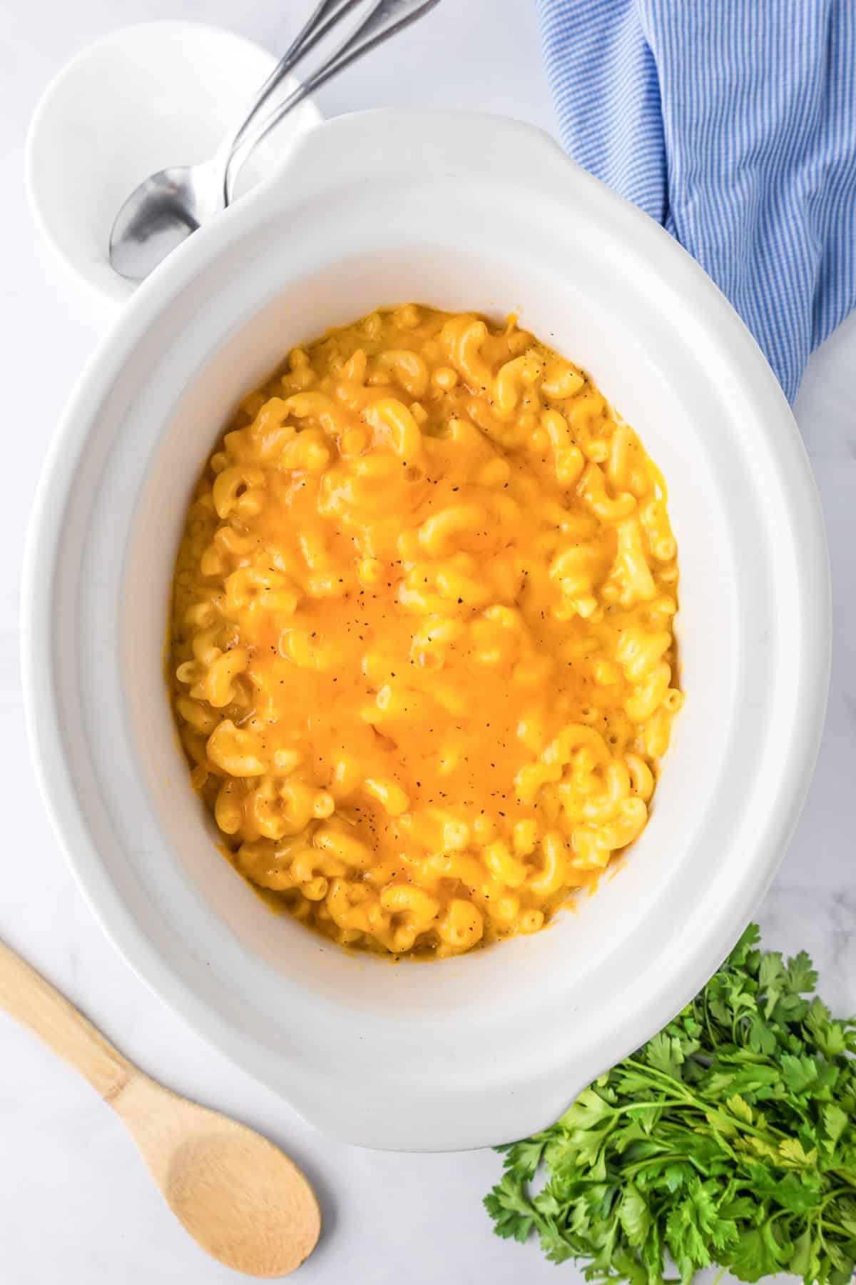 A slow cooker filled with creamy slow cooker mac and cheese sits on a white surface, surrounded by a wooden spoon, fresh parsley, and a blue striped cloth.