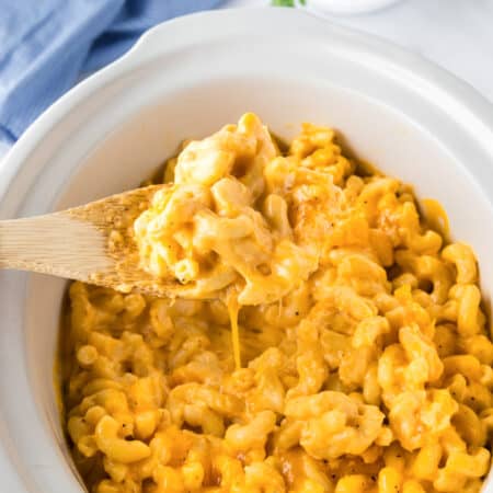 A wooden spoon lifts a serving of creamy slow cooker mac and cheese from a white slow cooker filled with the delicious dish.