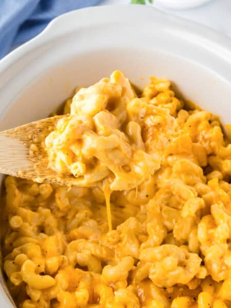 A wooden spoon lifts a serving of creamy slow cooker mac and cheese from a white slow cooker filled with the delicious dish.