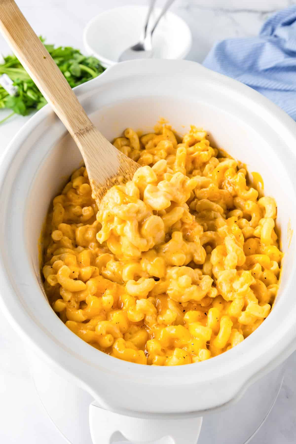 Slow cooker mac and cheese being stirred with a wooden spoon in a white slow cooker; chopped green onions and bowls sit in the background.