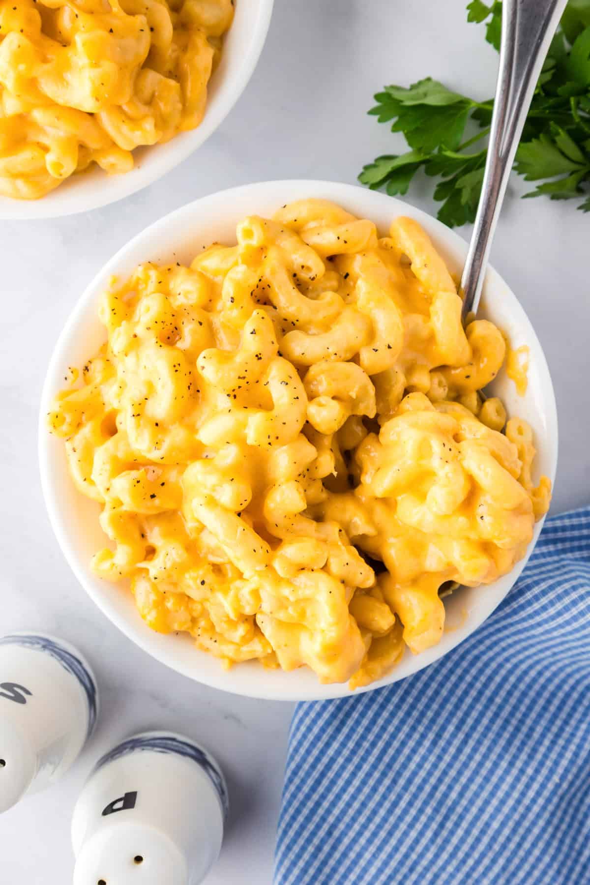 A bowl of creamy slow cooker mac and cheese with black pepper on top, placed next to a sprig of parsley, salt and pepper shakers, and a blue striped napkin.
