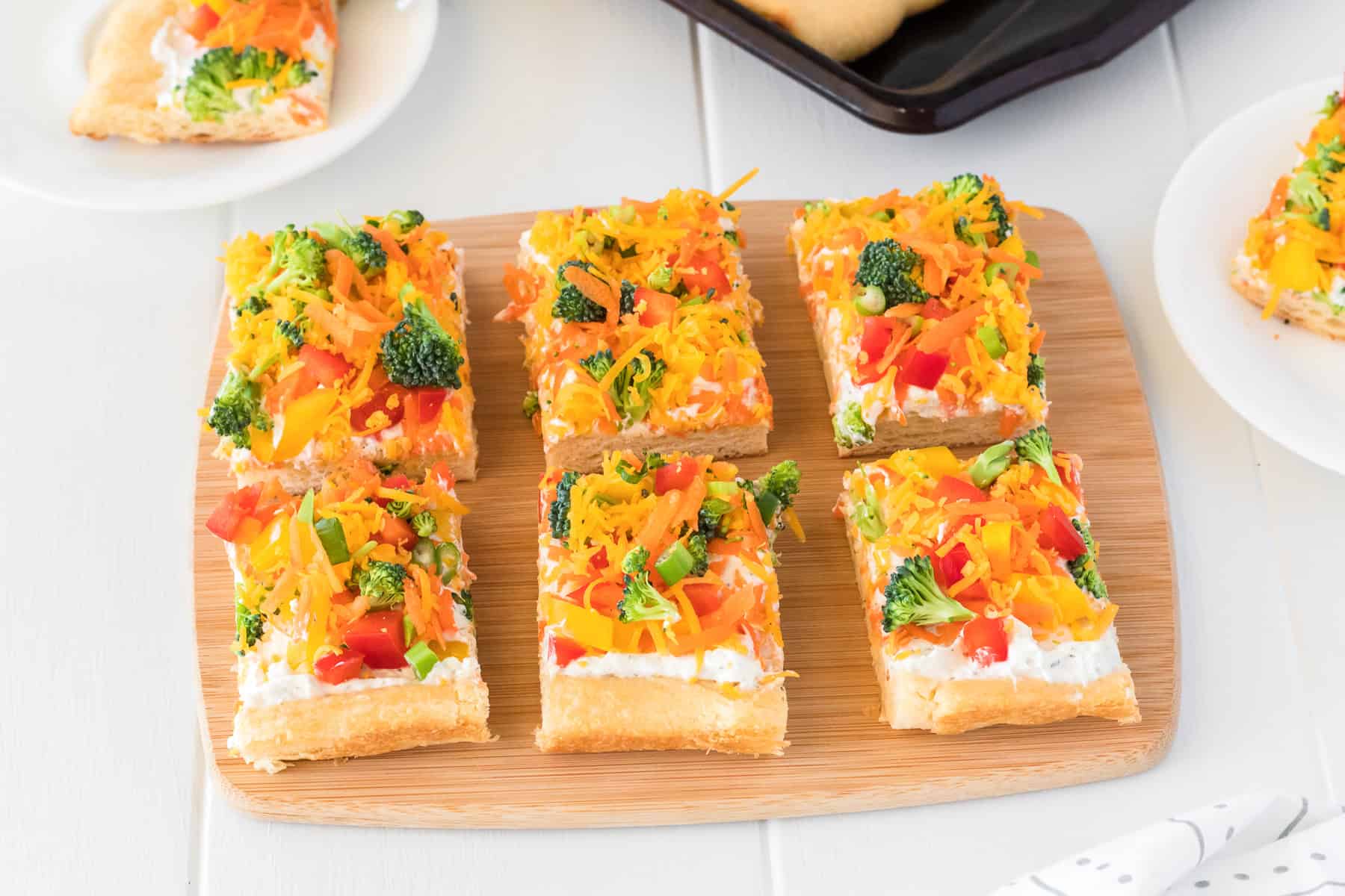 Six rectangular pieces of cold veggie pizza with a cream cheese base, topped with chopped broccoli, bell peppers, and shredded cheese, served on a wooden board.