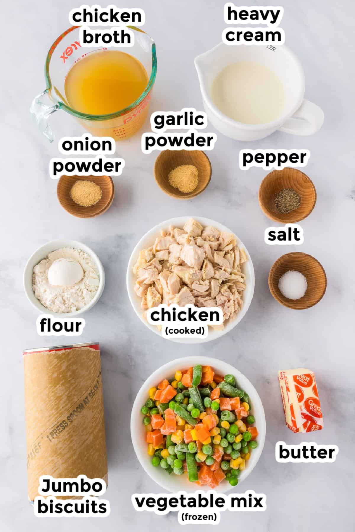 Overhead view of ingredients for a biscuit chicken pot pie: chicken broth, heavy cream, cooked chicken, frozen vegetable mix, jumbo biscuits, butter, flour, onion powder, garlic powder, salt, and pepper.