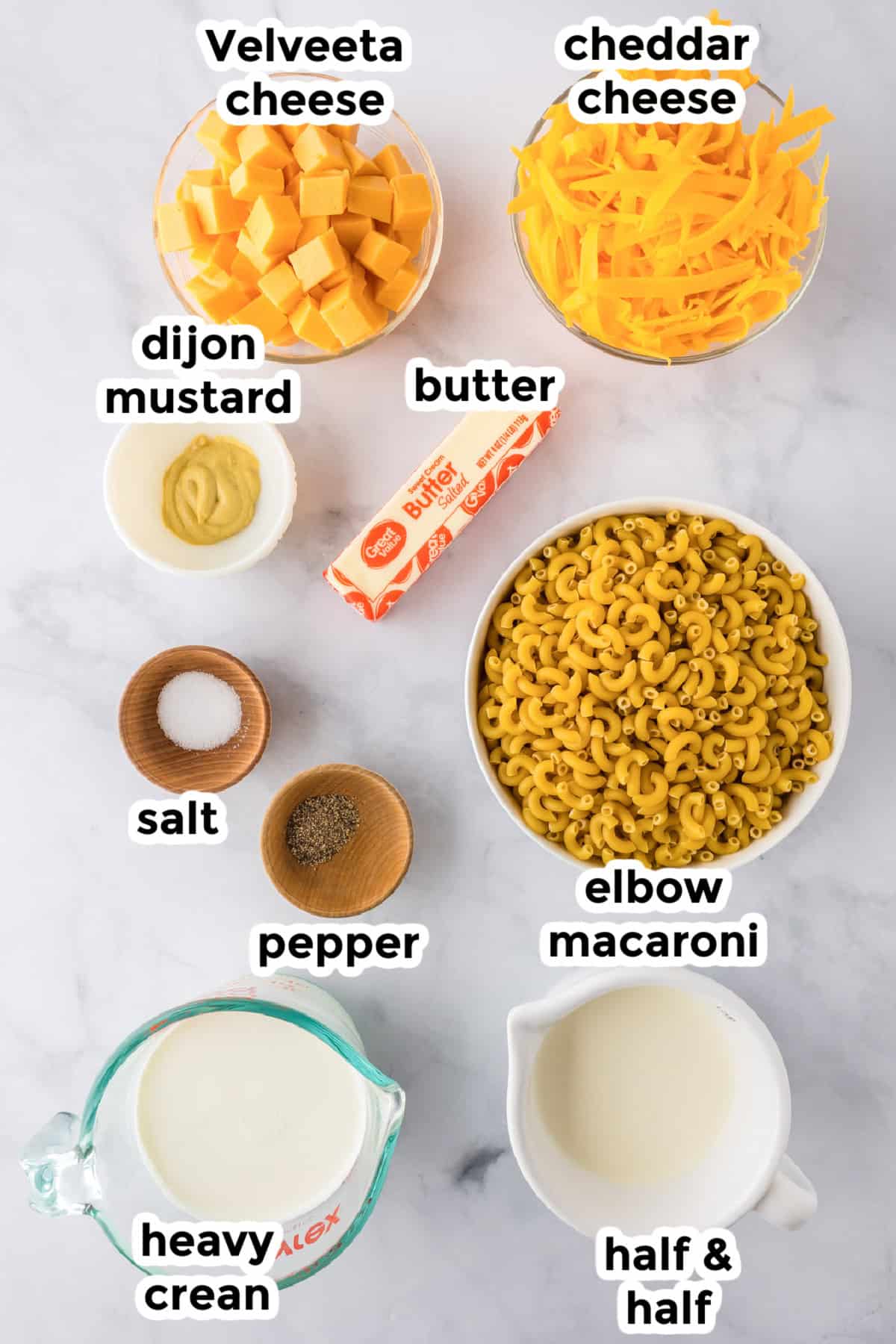 Ingredients for slow cooker mac and cheese are arranged in bowls on a counter, including cubes of Velveeta, shredded cheddar, elbow macaroni, butter, heavy cream, half & half, dijon mustard, salt, and pepper.