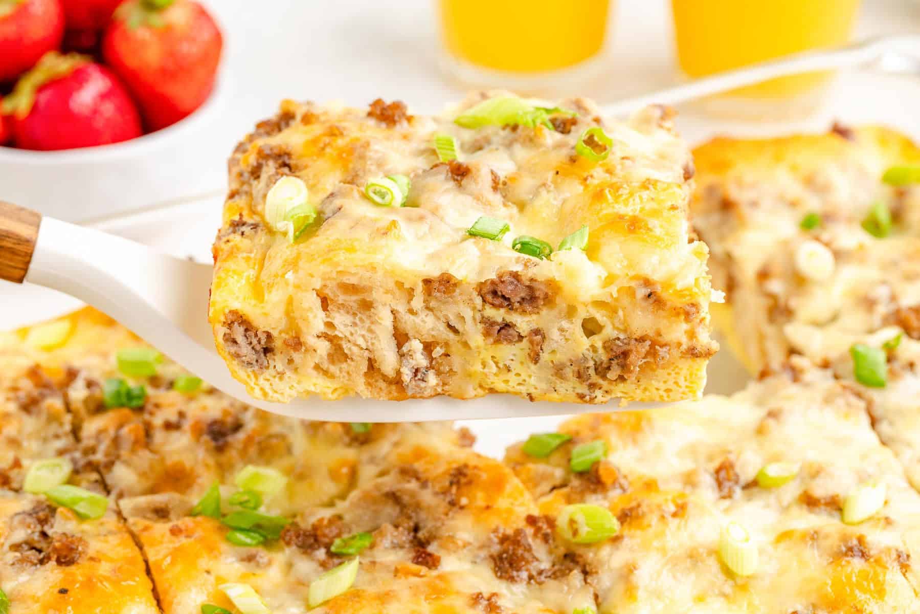 A spatula holds a slice of cheesy breakfast biscuit casserole with ground meat and green onions; strawberries and orange juice are in the background.