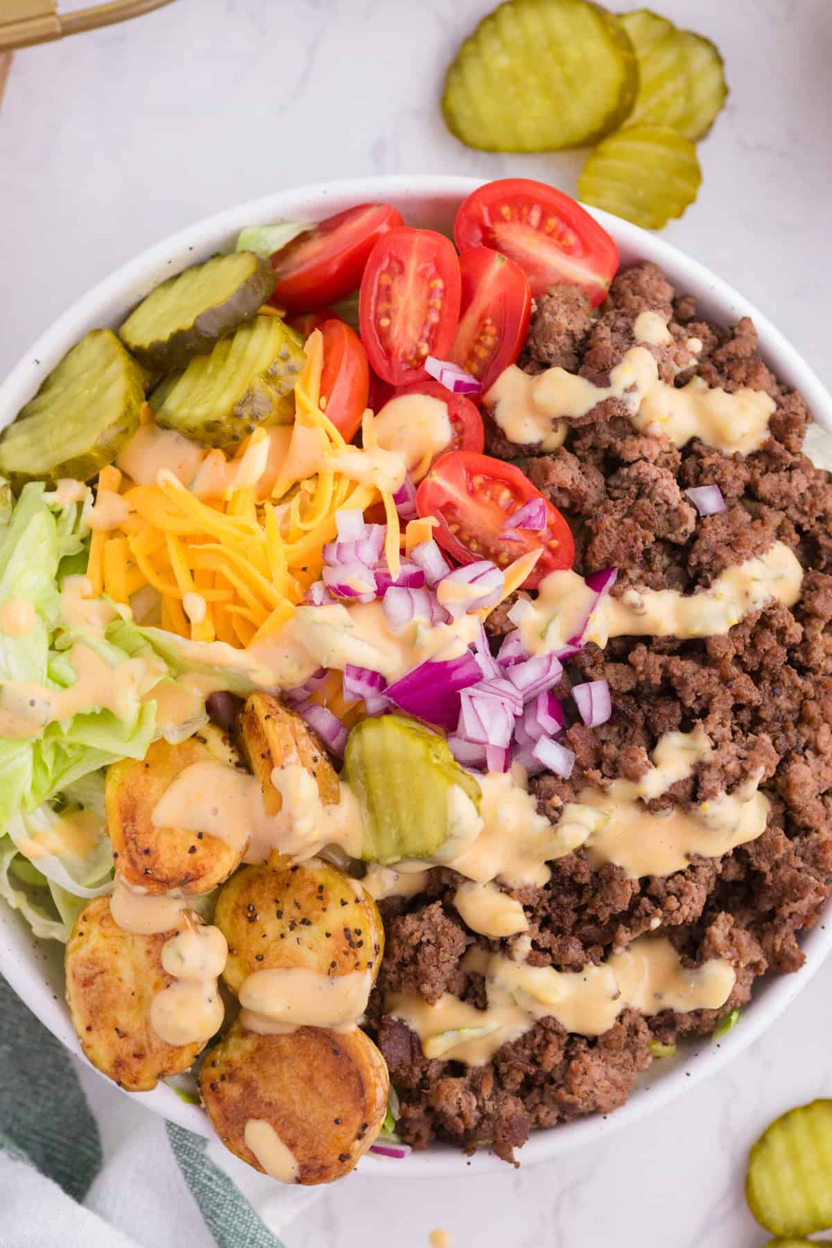 A burger bowl containing ground beef, sliced pickles, cherry tomatoes, shredded cheddar cheese, lettuce, diced red onion, roasted potatoes, and drizzled with a creamy sauce.