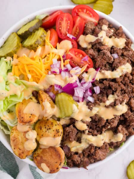 A burger bowl with ground beef, cherry tomatoes, pickles, shredded lettuce, shredded cheese, diced red onion, roasted potatoes, and dressing drizzled on top.