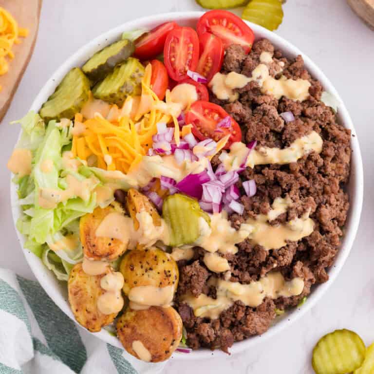 A burger bowl with ground beef, cherry tomatoes, pickles, shredded lettuce, shredded cheese, diced red onion, roasted potatoes, and dressing drizzled on top.