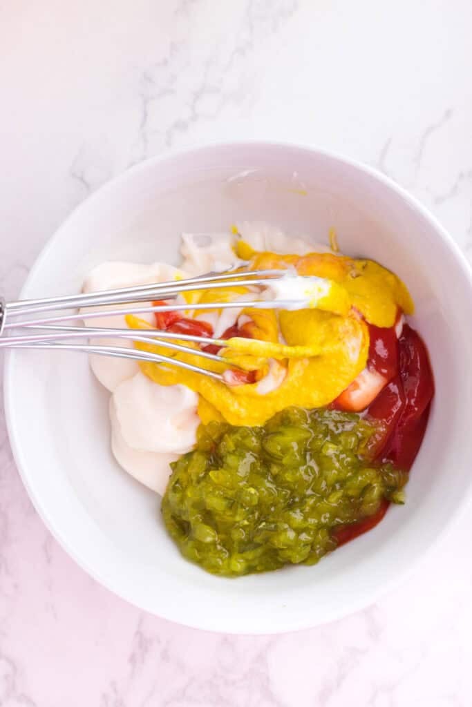 A white bowl containing mayonnaise, ketchup, mustard, and relish with a metal whisk on a white marble surface being mixed together to make burger sauce for a burger bowl.