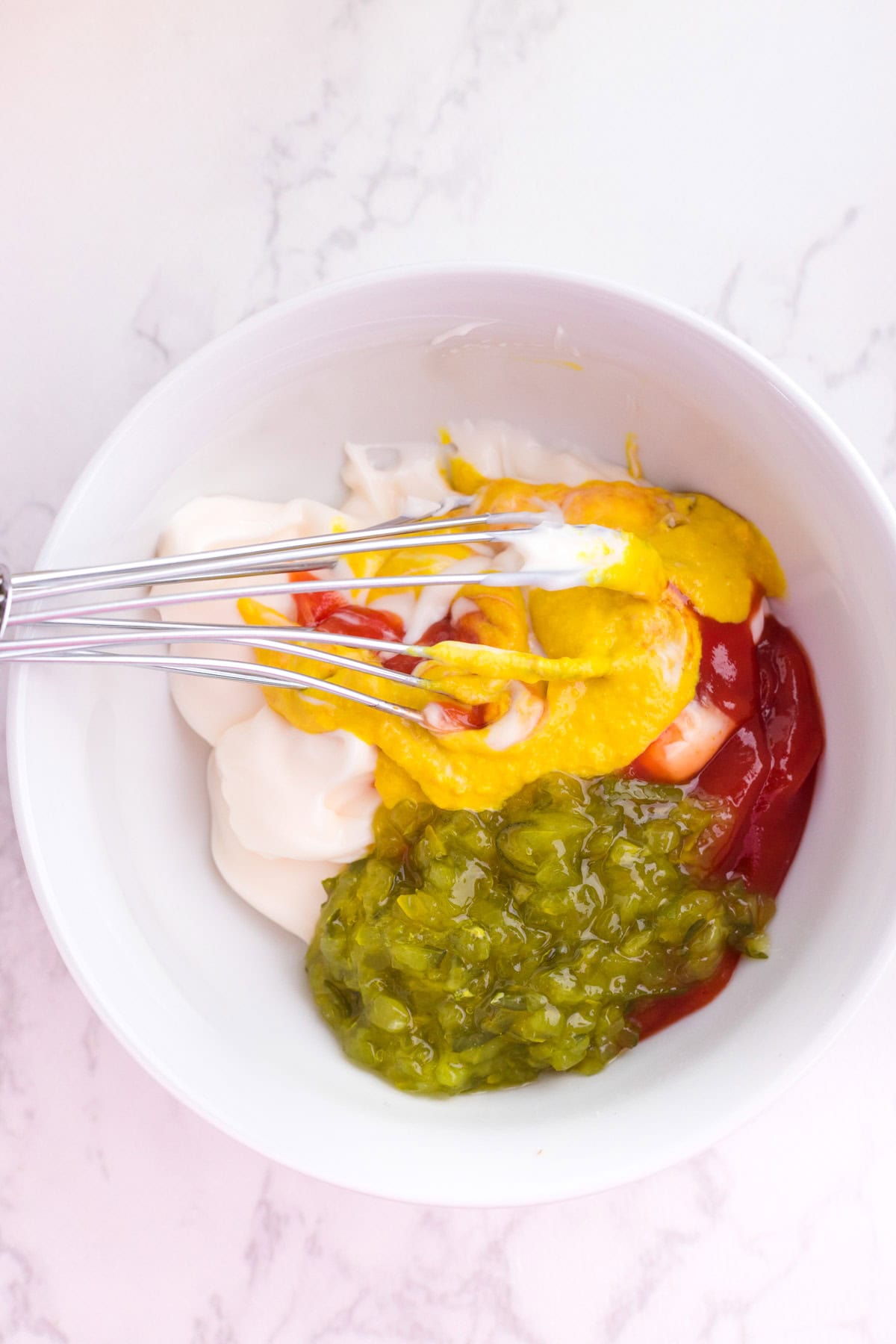 A white bowl containing mayonnaise, ketchup, mustard, and relish with a metal whisk on a white marble surface being mixed together to make burger sauce for a burger bowl.
