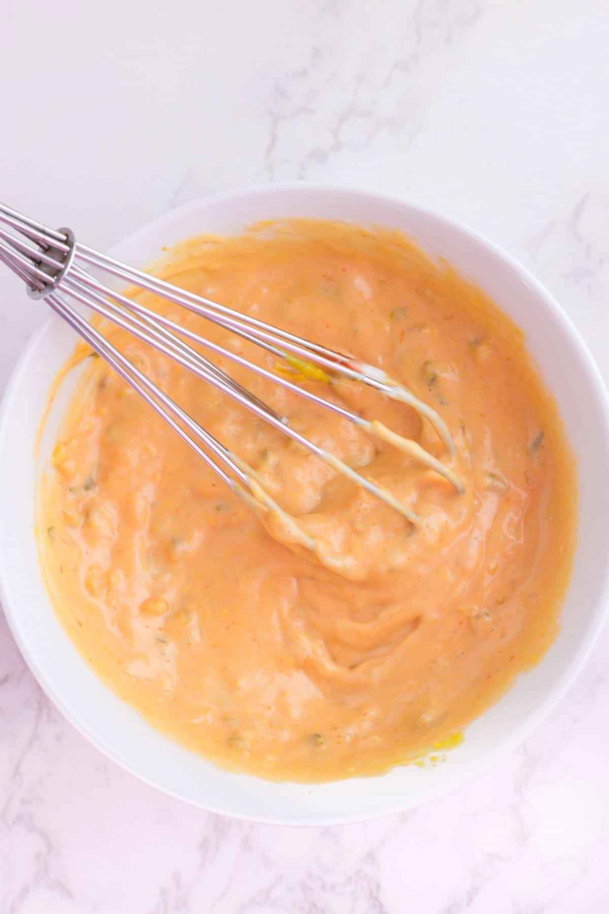 A metal whisk rests in a white bowl filled with a creamy, orange-colored burger sauce for a burger bowl.