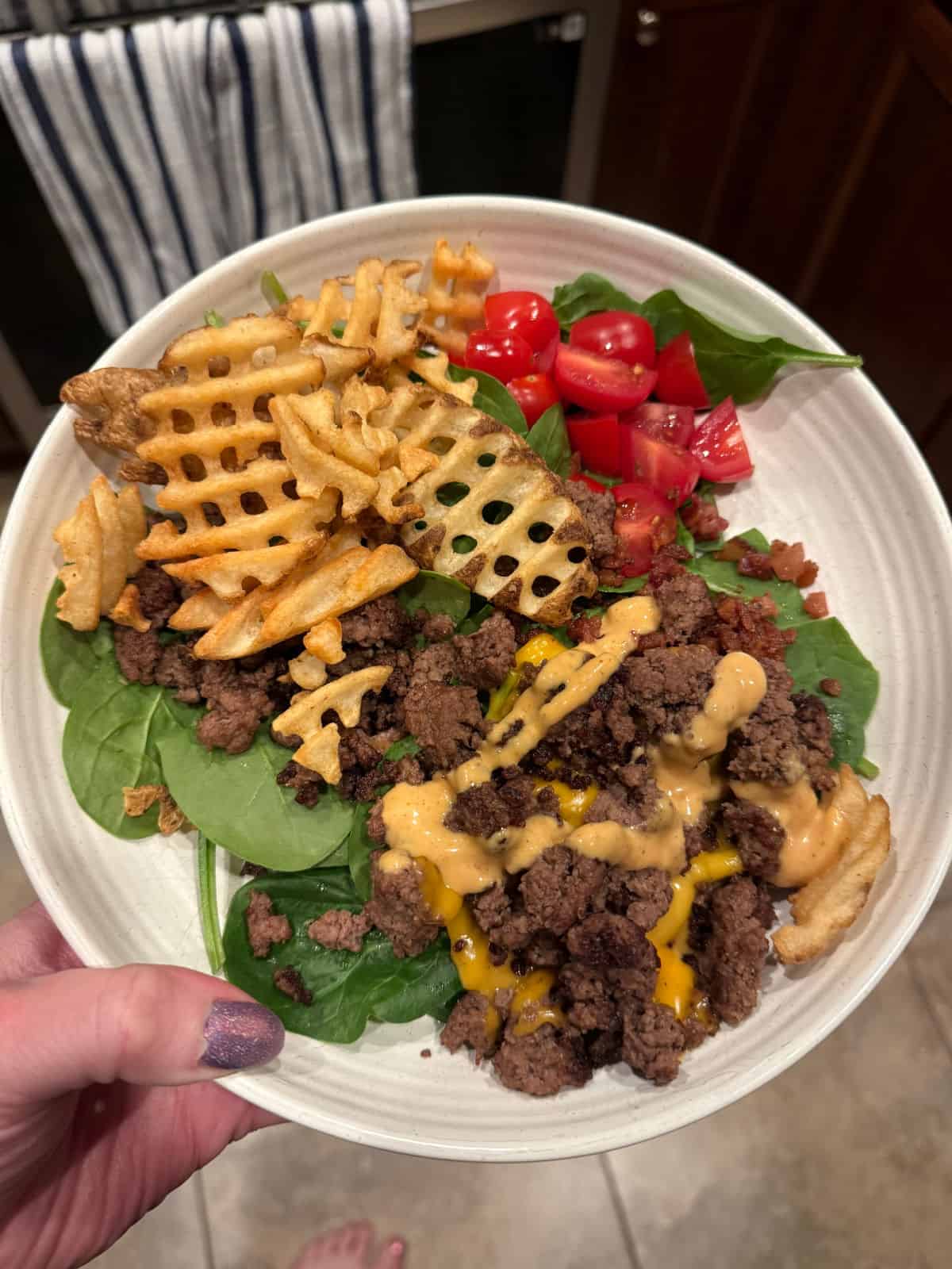 A burger bowl featuring ground beef with cheese sauce, waffle fries, fresh spinach, and sliced cherry tomatoes, all served in a bowl held above a tiled floor.