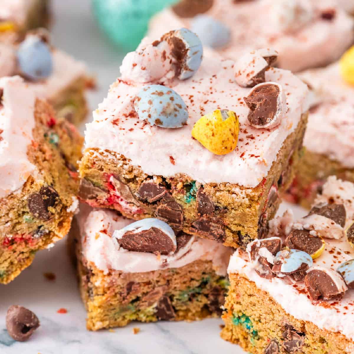 Frosted mini egg cookie bars topped with colorful chocolate candy eggs and chocolate pieces, stacked on a marble surface.
