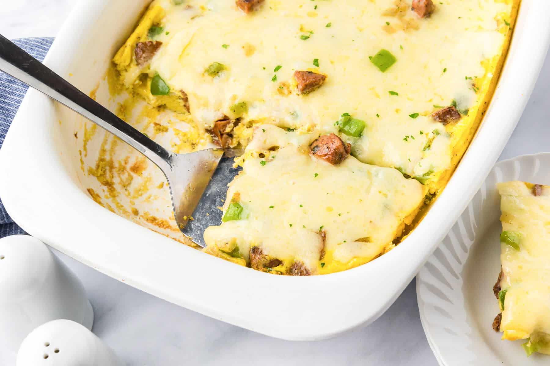 A chicken sausage egg bake breakfast casserole topped with melted cheese and green bell peppers in a white dish, with a metal spatula and a serving on a plate nearby.