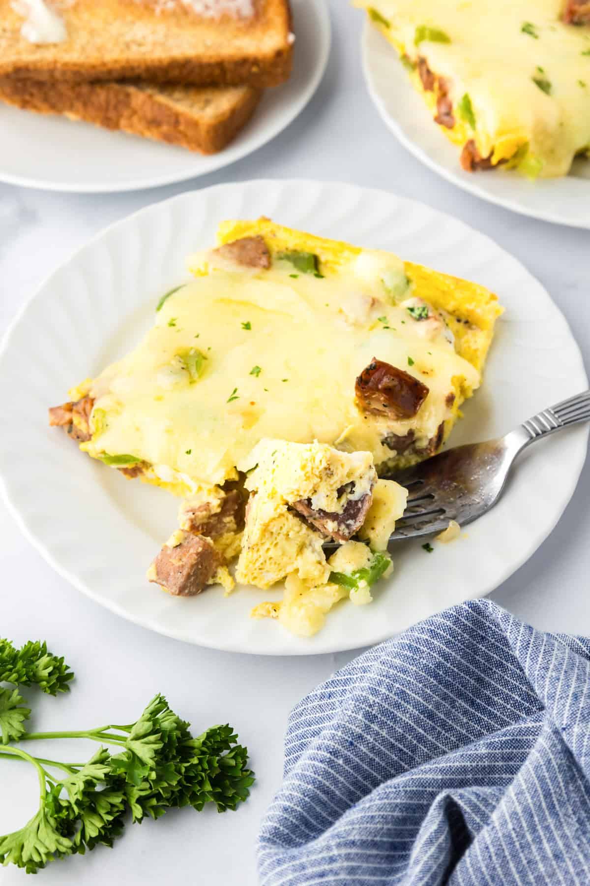 A slice of chicken sausage egg bake breakfast casserole on a white plate with a fork, garnished with parsley, with toast and a blue striped napkin nearby.