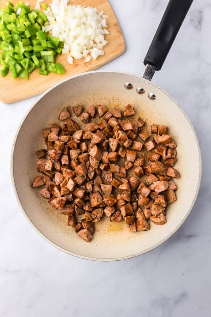 Diced sausage cooking in a skillet with chopped green bell pepper and onion on a cutting board in the background.