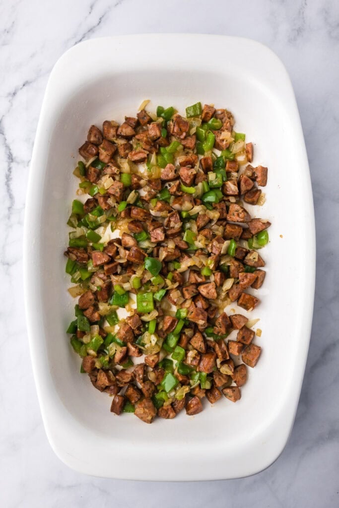 Diced chicken sausage and green bell peppers spread evenly in a white baking dish on a marble surface, ready for a delicious egg bake breakfast casserole.