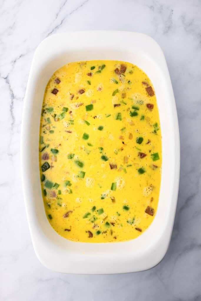 A rectangular white baking dish filled with a raw egg mixture containing chopped vegetables and possibly bacon, on a marble surface.