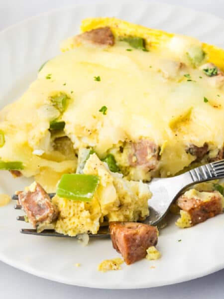 A slice of cheesy chicken sausage breakfast casserole with eggs and green bell peppers on a white plate, partially eaten with a fork.