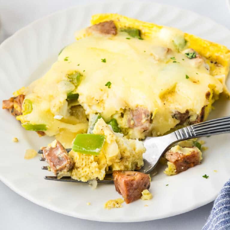 A slice of cheesy chicken sausage breakfast casserole with eggs and green bell peppers on a white plate, partially eaten with a fork.