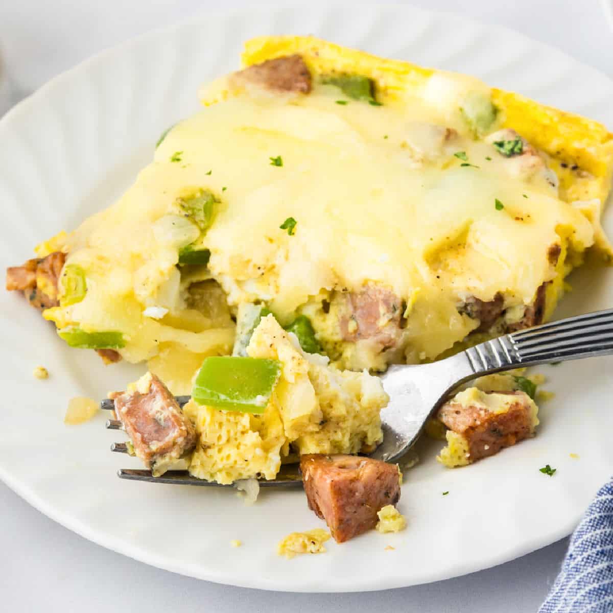 A slice of cheesy chicken sausage breakfast casserole with eggs and green bell peppers on a white plate, partially eaten with a fork.