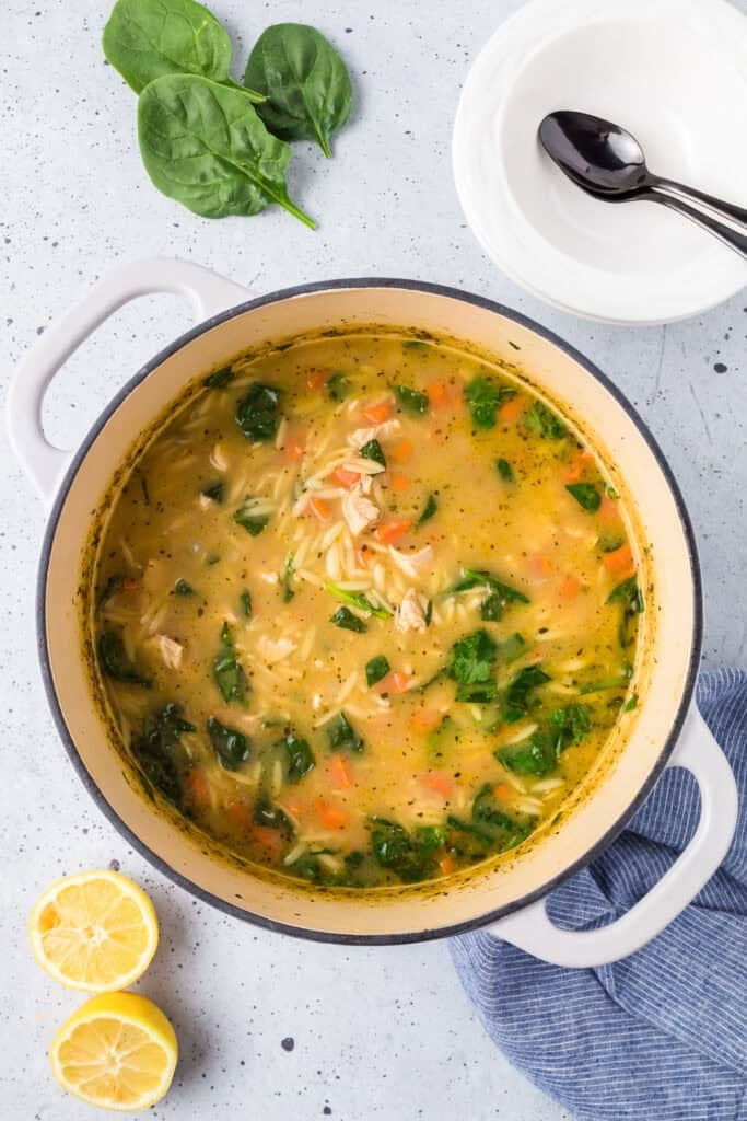 A pot of lemon chicken orzo soup with spinach and carrots sits on a counter next to lemon halves, fresh spinach leaves, and stacked white bowls with spoons.