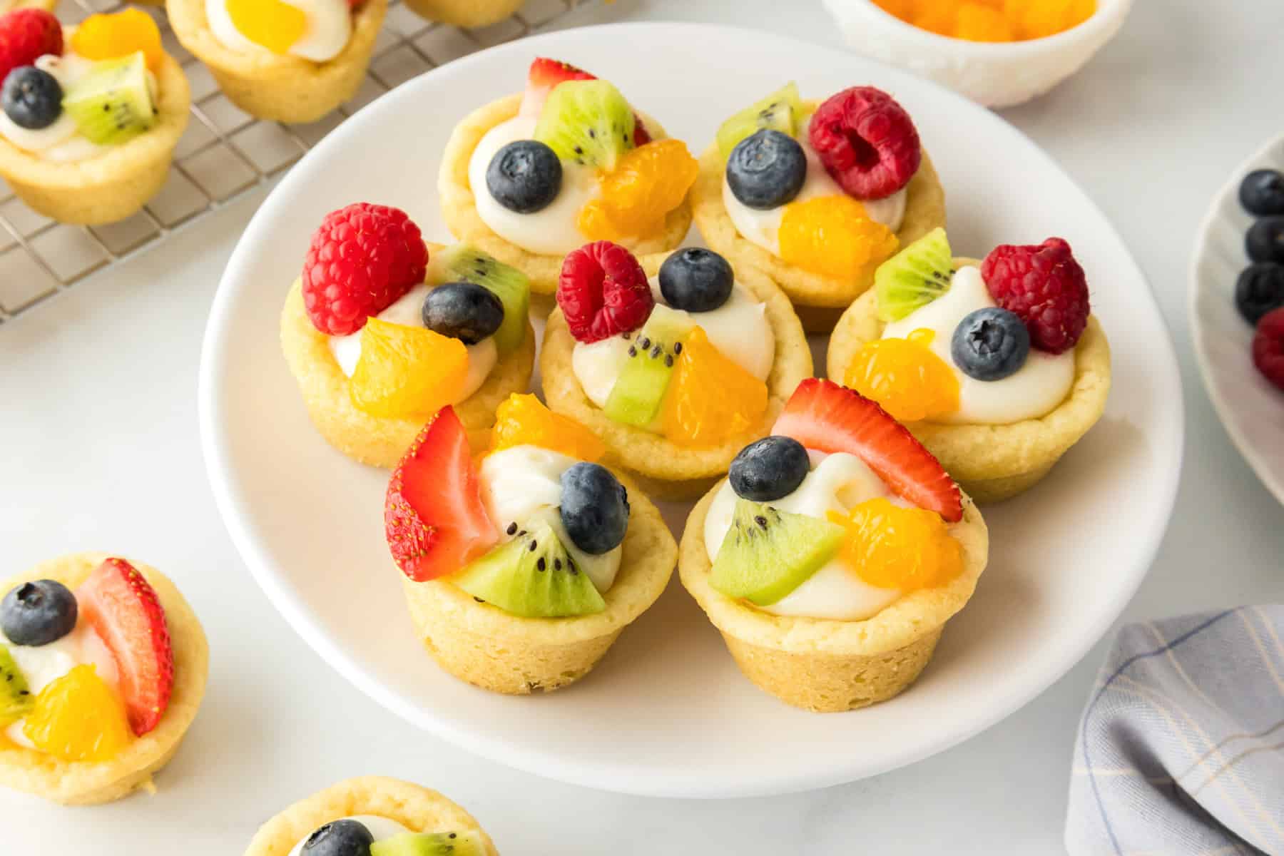 A plate of mini fruit tarts made in sugar cookie cups with vanilla cream filling, topped with strawberries, kiwis, blueberries, raspberries, and mandarin orange segments.