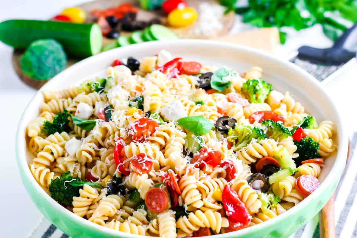 A bowl of Italian Pasta Salad with rotini, cherry tomatoes, broccoli, olives, mozzarella chunks, and grated cheese, with fresh vegetables visible in the background.