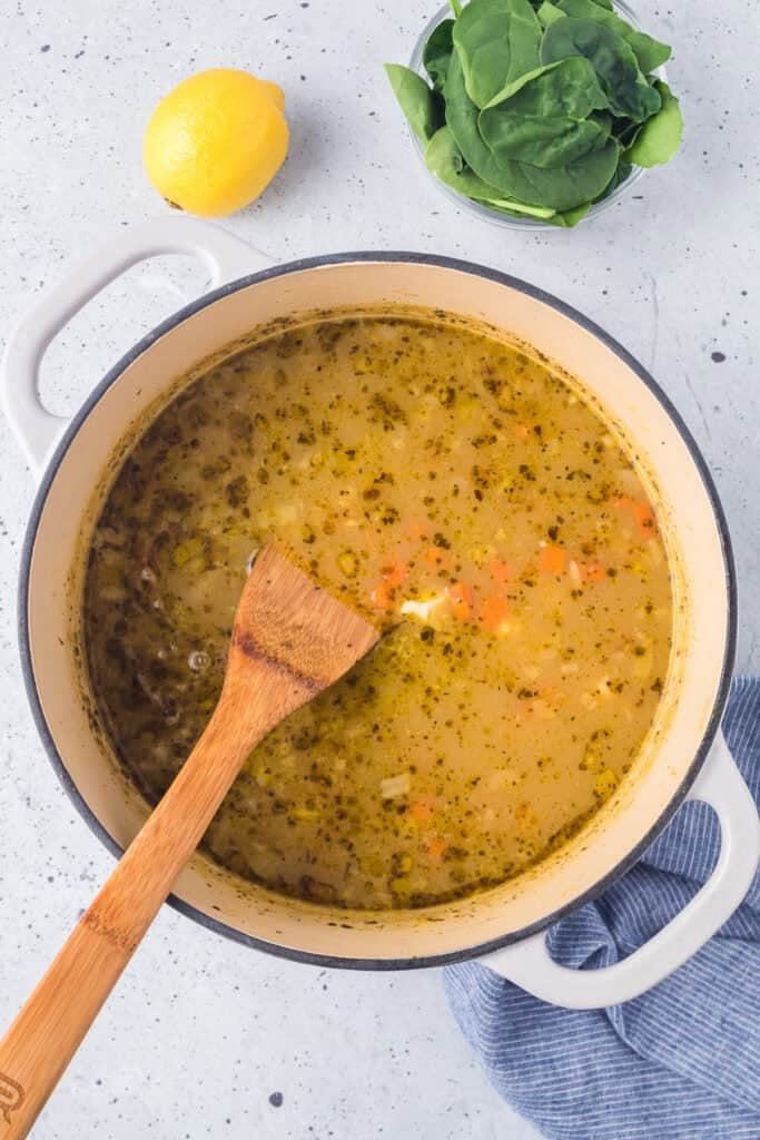 A pot of lemon chicken orzo soup with a wooden spoon stirring, next to a lemon, a bowl of spinach, and a blue cloth on a light countertop.