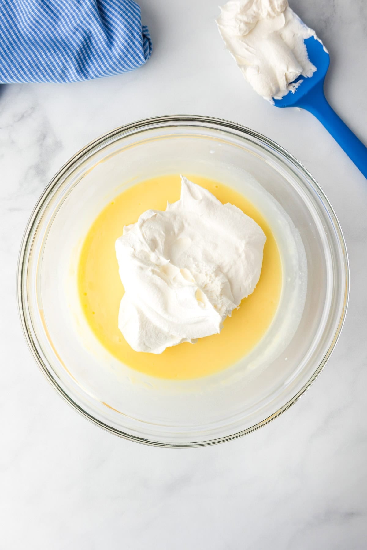 A glass bowl contains cheesecake pudding being mixed with Cool Whip for a layer of lemon eclair icebox cake.