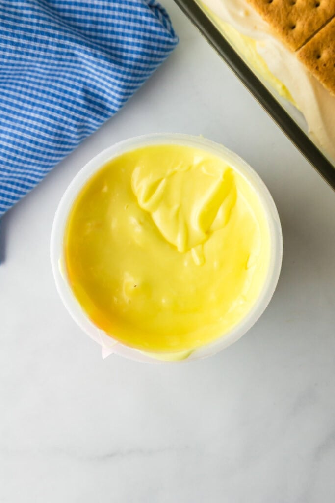 A container filled with lemon frosting after warming sits next to a casserole dish full of lemon eclair icebox cake.