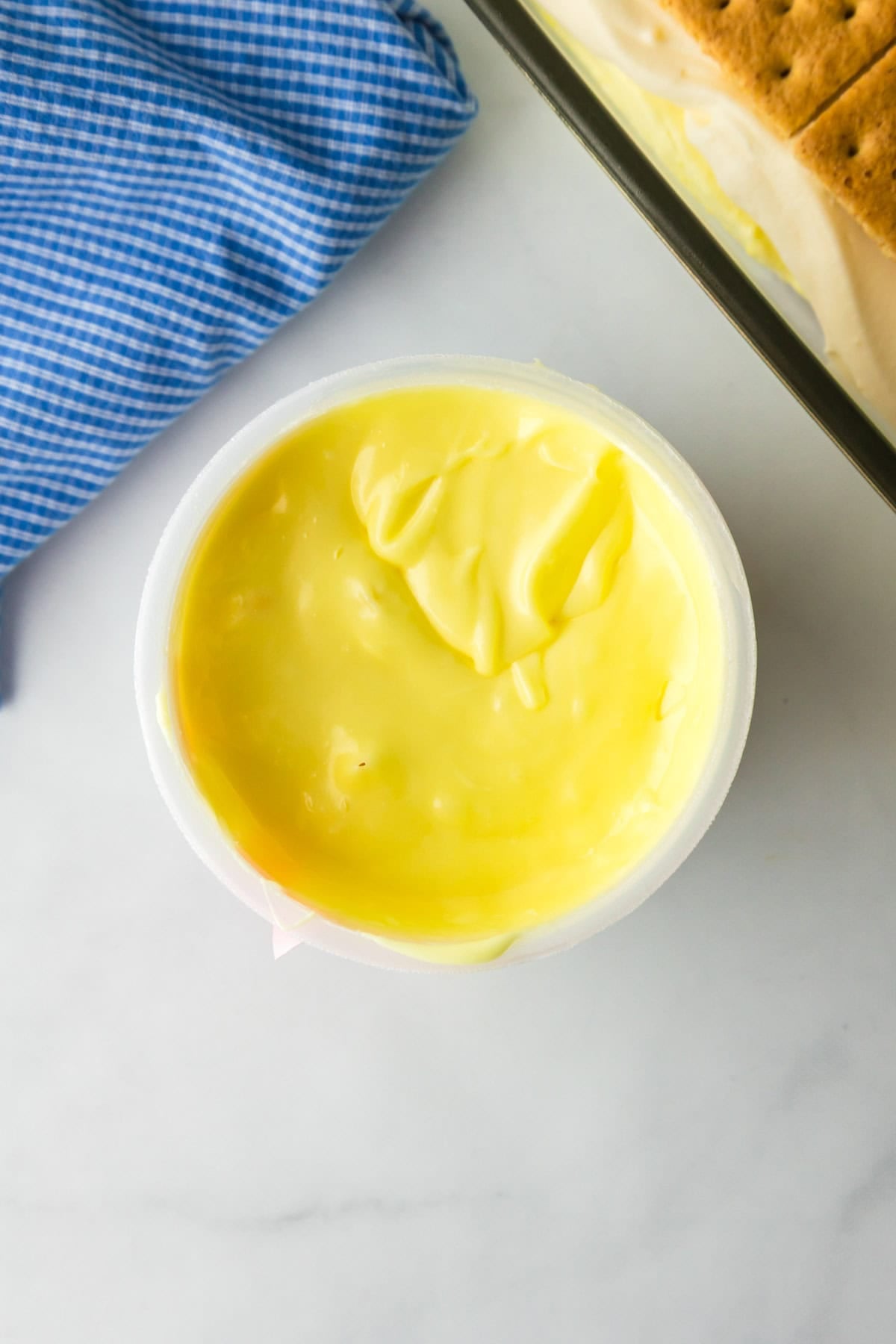 A container filled with lemon frosting after warming sits next to a casserole dish full of lemon eclair icebox cake.