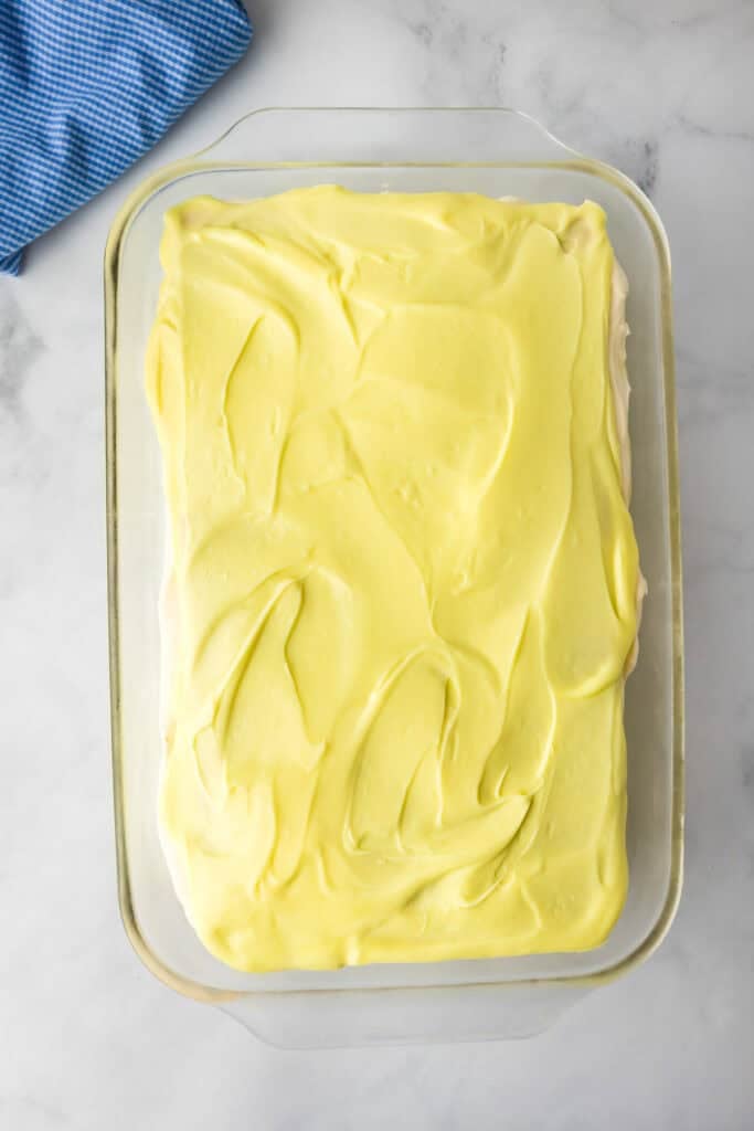 A glass baking dish filled with a finished lemon eclair icebox cake, topped with a smooth layer of yellow frosting, set on a white counter.